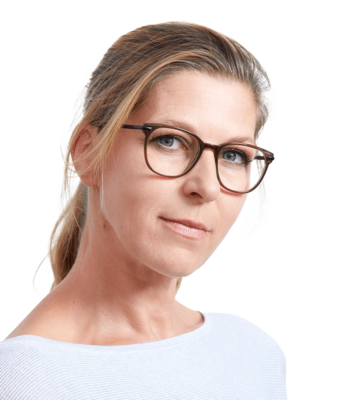 Eine Frau mit blondem Haar, die eine Brille und ein helles Oberteil trägt, blickt ruhig in die Kamera vor einem schlichten weißen Hintergrund.