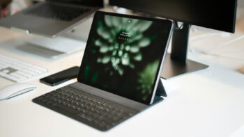 Ein Tablet mit Tastaturgehäuse zeigt einen Sperrbildschirm mit einer Passworteingabe an und steht auf einem weißen Schreibtisch neben einem Monitor, einer drahtlosen Maus, einer Tastatur und einem Smartphone.