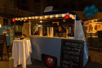 Ein gemütlicher Foodtruck mit Lichterketten serviert abends neben warmen Getränken auch SPiNNpunsch. Im Truck bereitet eine Person ein Getränk zu, während auf einer Tafel leckere Gerichte aufgelistet sind. In der Nähe bietet ein kleiner Tisch Erfrischungen an, während blaue Luftballons der Szene eine festliche Note verleihen.