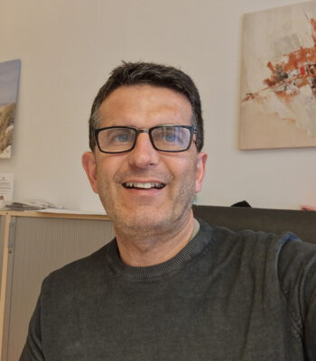 Ein Mann mit kurzen dunklen Haaren, Brille und dunklem Pullover sitzt im Haus und lächelt in die Kamera. Hinter ihm hängt ein abstraktes Kunstwerk an der Wand, das sein Interesse an Design und die Auswirkungen des Barrierefreiheitsgesetzes unterstreicht.