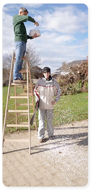 Eine Person steht auf einer Holzleiter und schüttet Konfetti auf eine andere Person, die Schutzkleidung trägt und auf einem gepflasterten Weg steht. Die Szenerie unterstreicht die Barrierefreiheitsanforderungen des Barrierefreiheitsgesetzes, mit Gras und Häusern im Hintergrund.