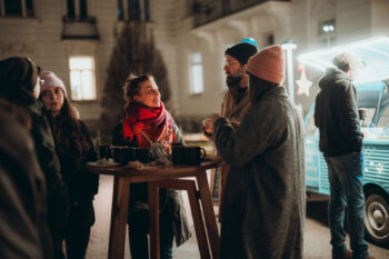Eine kleine Gruppe winterlich gekleideter Menschen steht nachts im Freien an einem hohen Holztisch, plaudert und schlürft SPiNNpunsch in der Nähe eines beleuchteten Foodtrucks. Die Szene sieht warm und festlich aus.