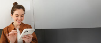 Eine lächelnde Frau mit braunem Haar in einem Dutt benutzt ein Smartphone mit einem Stift. Sie trägt ein braunes Hemd und steht vor einem schlichten hellgrauen Hintergrund.