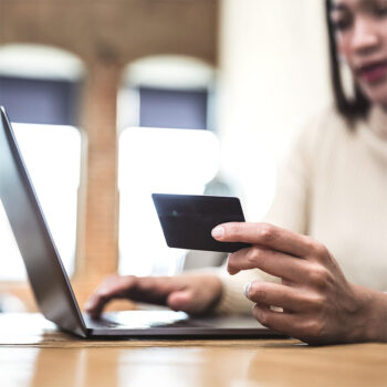 Eine Person hält eine Kreditkarte in der einen Hand, während sie mit der anderen Hand auf einem Laptop tippt, was auf Online-Shopping oder Online-Banking hindeutet. Der Hintergrund ist unscharf.