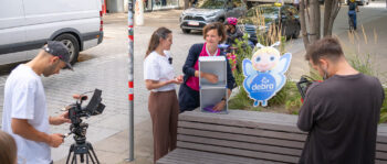 Zwei Frauen stehen an einem kleinen Schrank im Freien in der Nähe eines Debra-Schildes, während zwei Männer sie mit Kameras auf einem städtischen Bürgersteig filmen. Im Hintergrund sind Bäume, geparkte Fahrzeuge und eine Bank zu sehen.