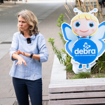 Eine Frau gestikuliert, während sie neben einem Cartoon-Schmetterlingsmaskottchen steht, das ein Schild mit der Aufschrift Debra hält, im Freien auf einer Straße in der Nähe eines großen Pflanzkübels mit Grünpflanzen.