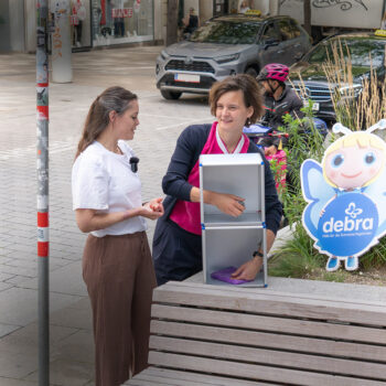 Zwei Frauen stehen in der Nähe eines Gehwegaufstellers mit einer Cartoon-Engelsfigur und dem Debra-Logo. Eine Frau legt einen lila Gegenstand in eine Metallbox, während die andere spricht. Im Hintergrund sind Radfahrer und städtische Landschaften zu sehen.