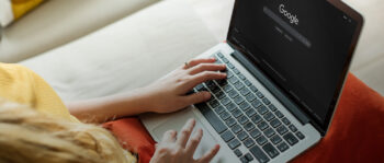 Eine Person mit einem Laptop, auf dem die Google-Homepage im dunklen Modus geöffnet ist, tippt auf der Tastatur, während sie auf einer Couch mit einem orangefarbenen Kissen sitzt.