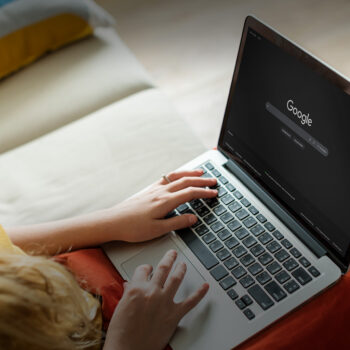 Eine Person, die einen Laptop benutzt, auf dessen Bildschirm die Startseite der Google-Suche angezeigt wird. Sie sitzt auf einem Sofa und tippt mit einer Hand auf der Tastatur, während die andere Hand auf dem Laptop ruht.